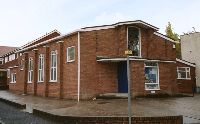 A small red-brick church building with a modern, angular roofline, tall white-framed windows along the side, and a blue door at the front entrance. A signpost stands on the pavement in front.