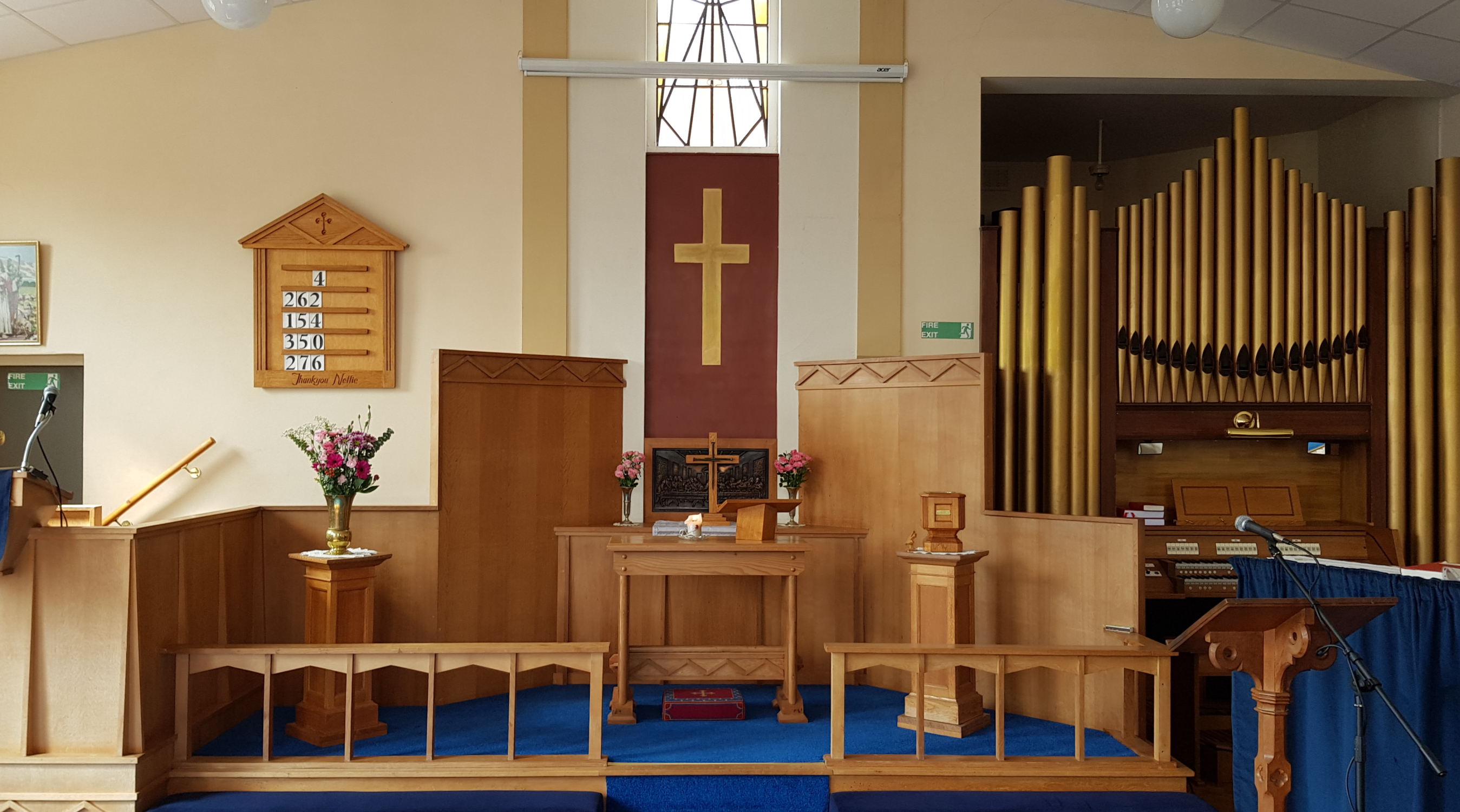The interior of the church showing the dais with a wooden communion table, a gold cross on a red backdrop, floral arrangements, hymn numbers displayed on a wooden board, an organ with pipes to the right, and a wooden pulpit with a microphone.